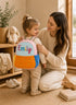 Mother helping a toddler wear a personalized orange mini backpack with custom name embroidery in a cozy nursery room