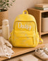 Personalized yellow quilted kids backpack with white custom name embroidery displayed on a wooden nursery shelf